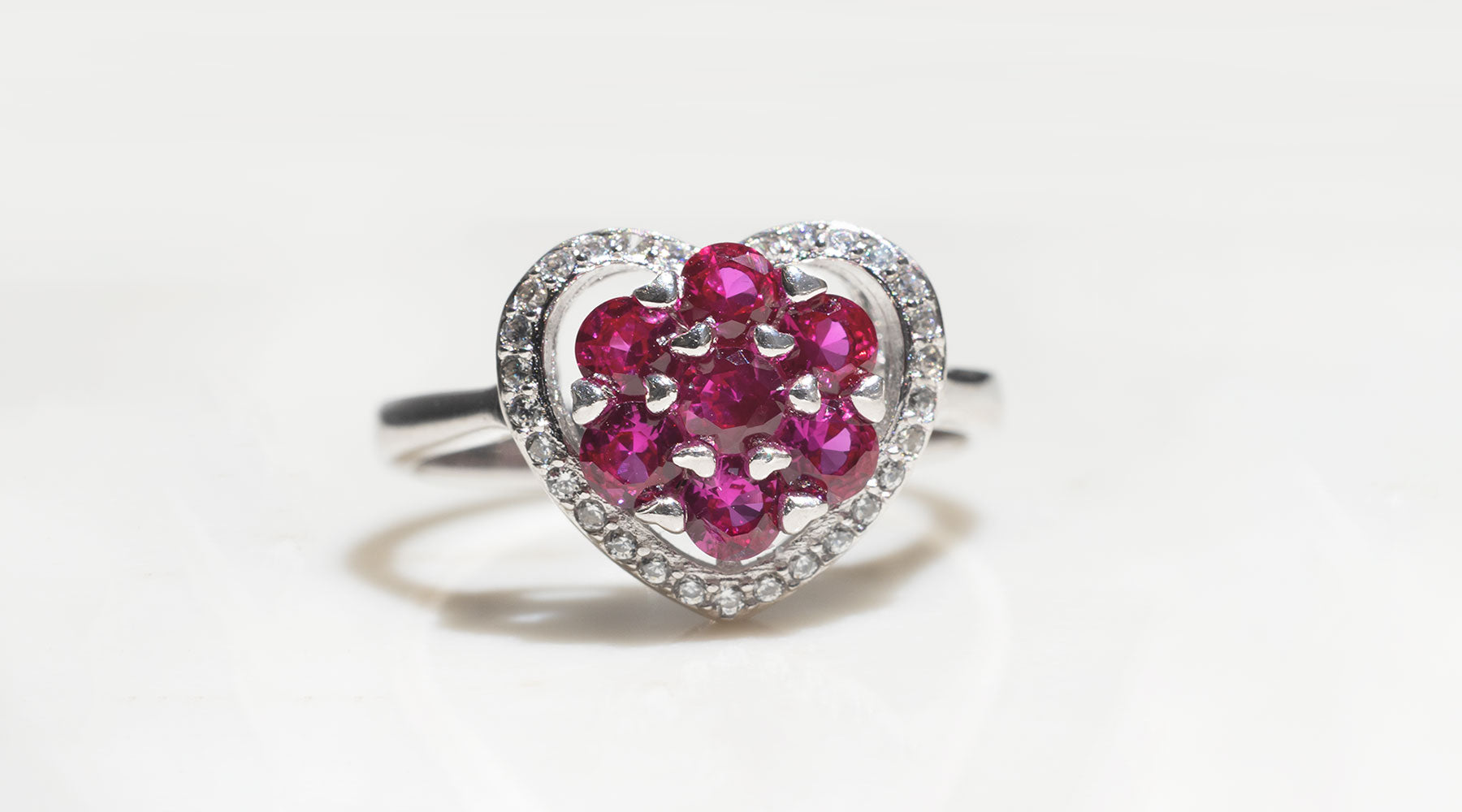 A white gold ring sits on a white surface.  The ring has a micro-grain set diamond heart-shaped setting.  In the center of the heart there is a cluster setting of seven round cut pigeon-blood rubies.  The rubies are held in place with heart shaped claws. 