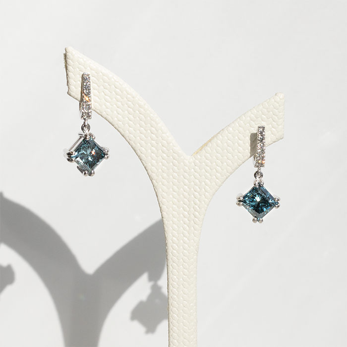 Pair of white gold drop earrings with square-cut blue diamonds and pavé diamond-set hooks displayed on a textured white stand.