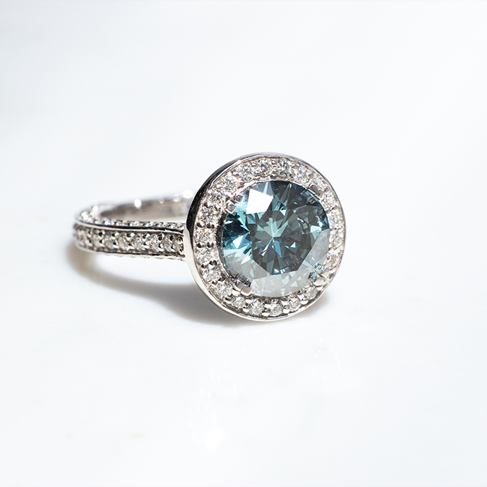 A white gold ring featuring a large round blue diamond with split-topped claws. There is a row of halo round brilliant cut lab grown diamonds on the outside of the blue diamond. the band features three rows of pave diamonds set on the outer edges.