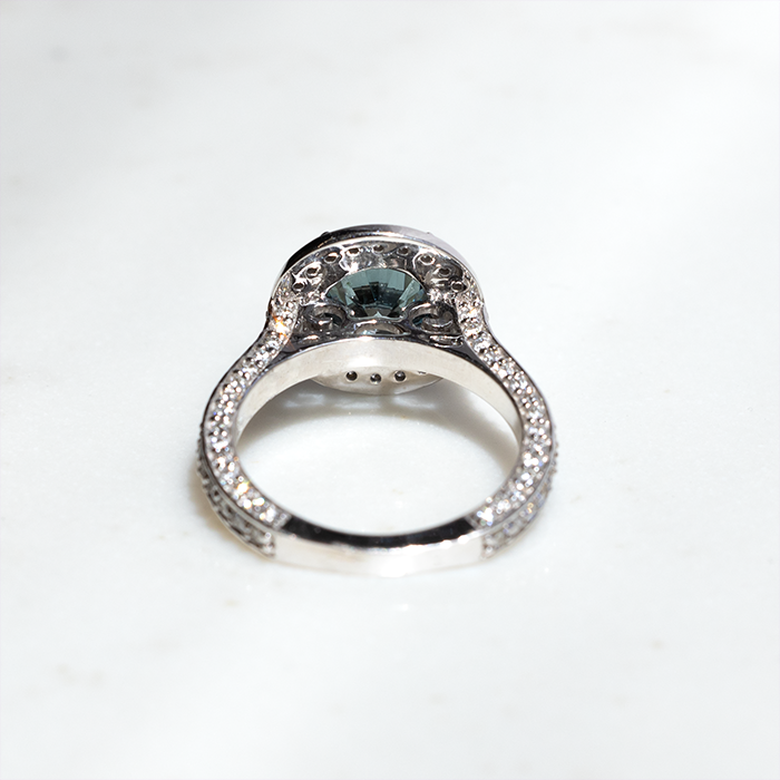 A white gold ring featuring a large round blue diamond with split-topped claws. There is a row of halo round brilliant cut lab grown diamonds on the outside of the blue diamond. the band features three rows of pave diamonds set on the outer edges.