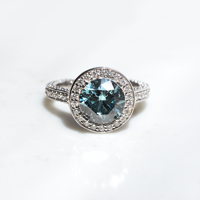 A white gold ring featuring a large round blue diamond with split-topped claws. There is a row of halo round brilliant cut lab grown diamonds on the outside of the blue diamond. the band features three rows of pave diamonds set on the outer edges.