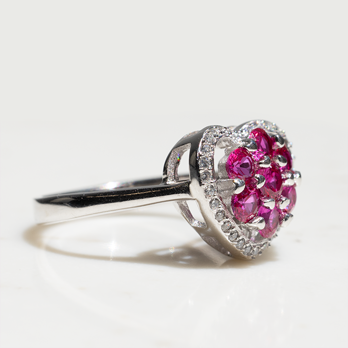 A heart-shaped ring with a cluster of red ruby stones in the center and white diamond stones around the halo, set in white gold or platinum.