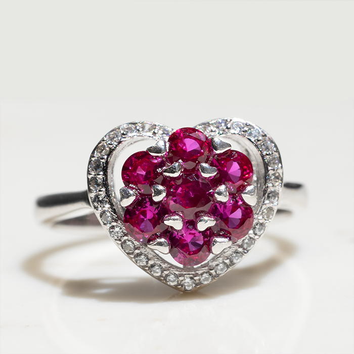 A heart-shaped ring with a cluster of red ruby stones in the center and white diamond stones around the halo, set in white gold or platinum.