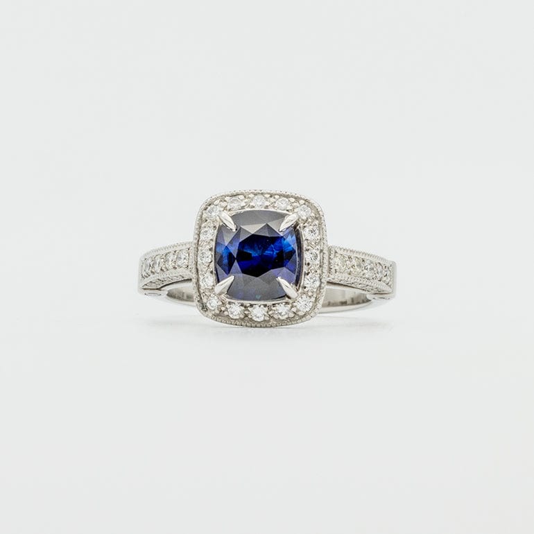 A Sapphire and Diamond Halo Engagement Ring with a cushion cut blue sapphire center stone, encircled by round diamonds set in a platinum band.
