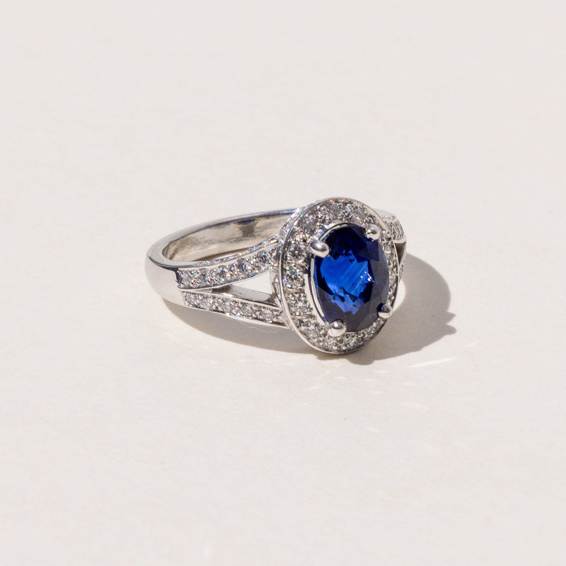 An oval blue sapphire halo ring with a split band and round brilliant cut diamonds, set in platinum.