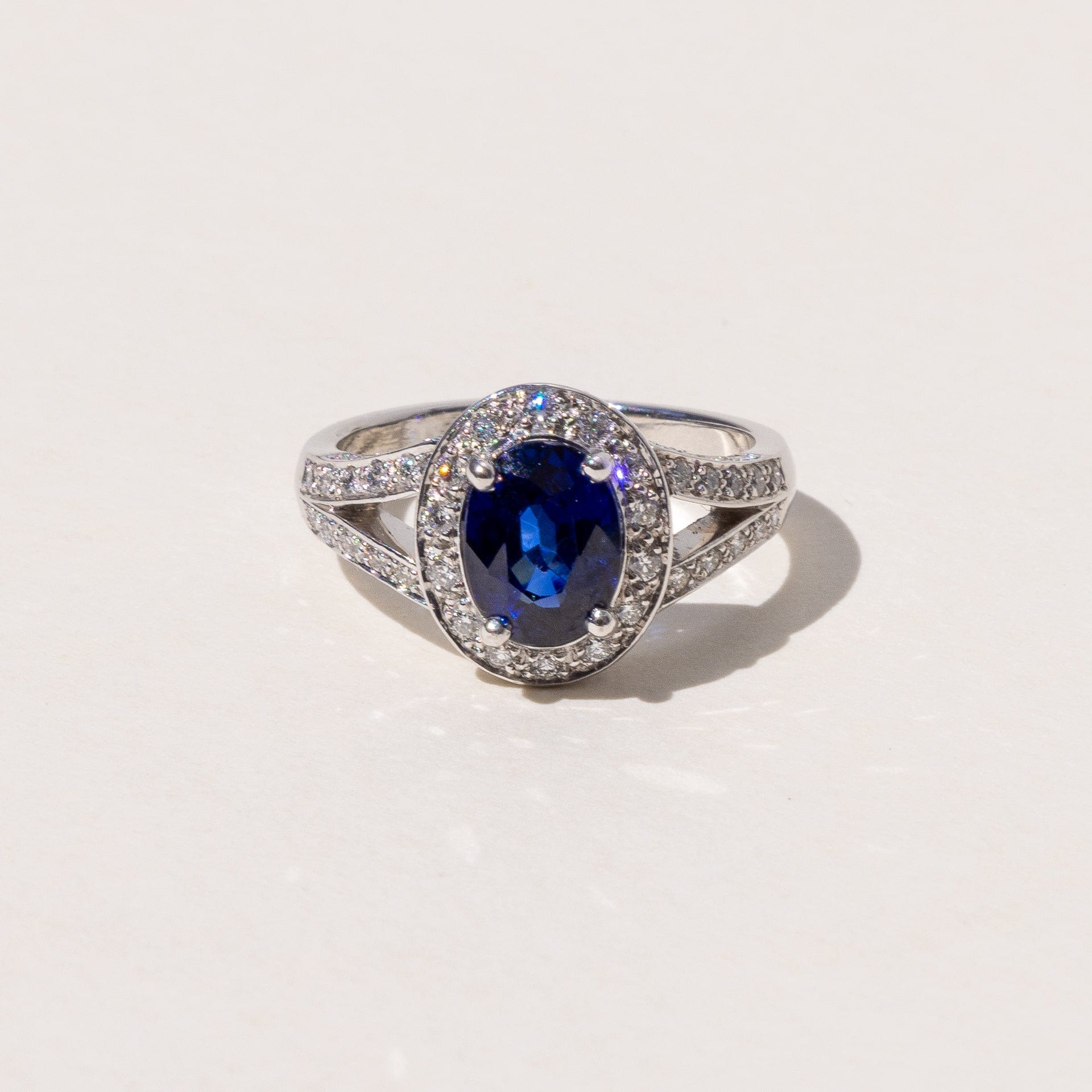 An oval blue sapphire halo ring with a split band and round brilliant cut diamonds, set in platinum.