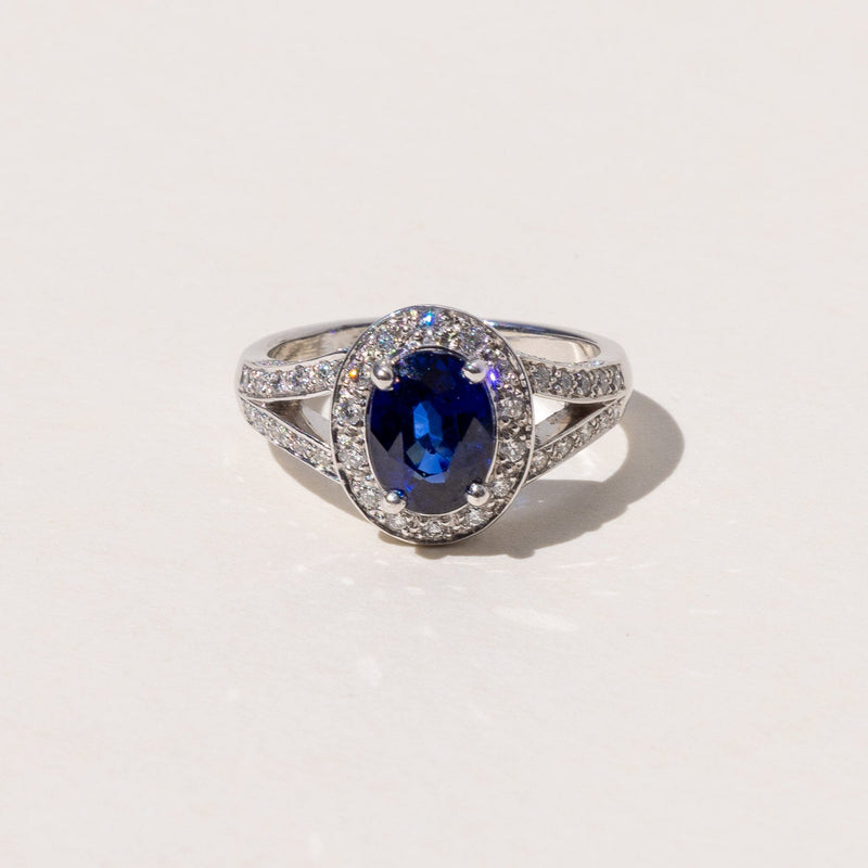 An oval blue sapphire halo ring with a split band and round brilliant cut diamonds, set in platinum.