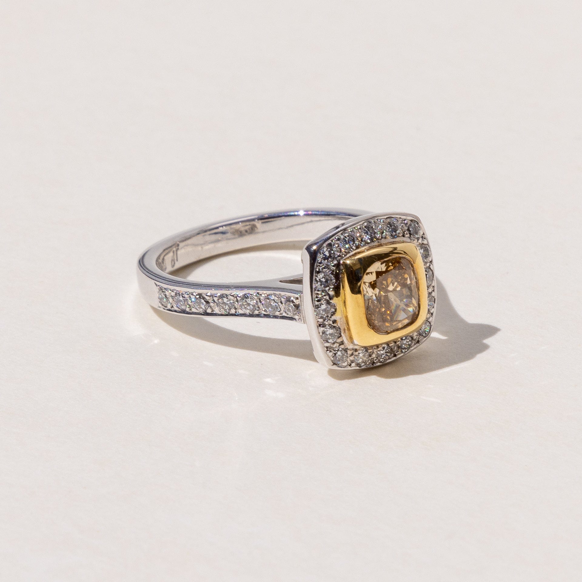 A square cushion cut cognac diamond halo ring with round brilliant cut diamonds around it, set in yellow and white gold.