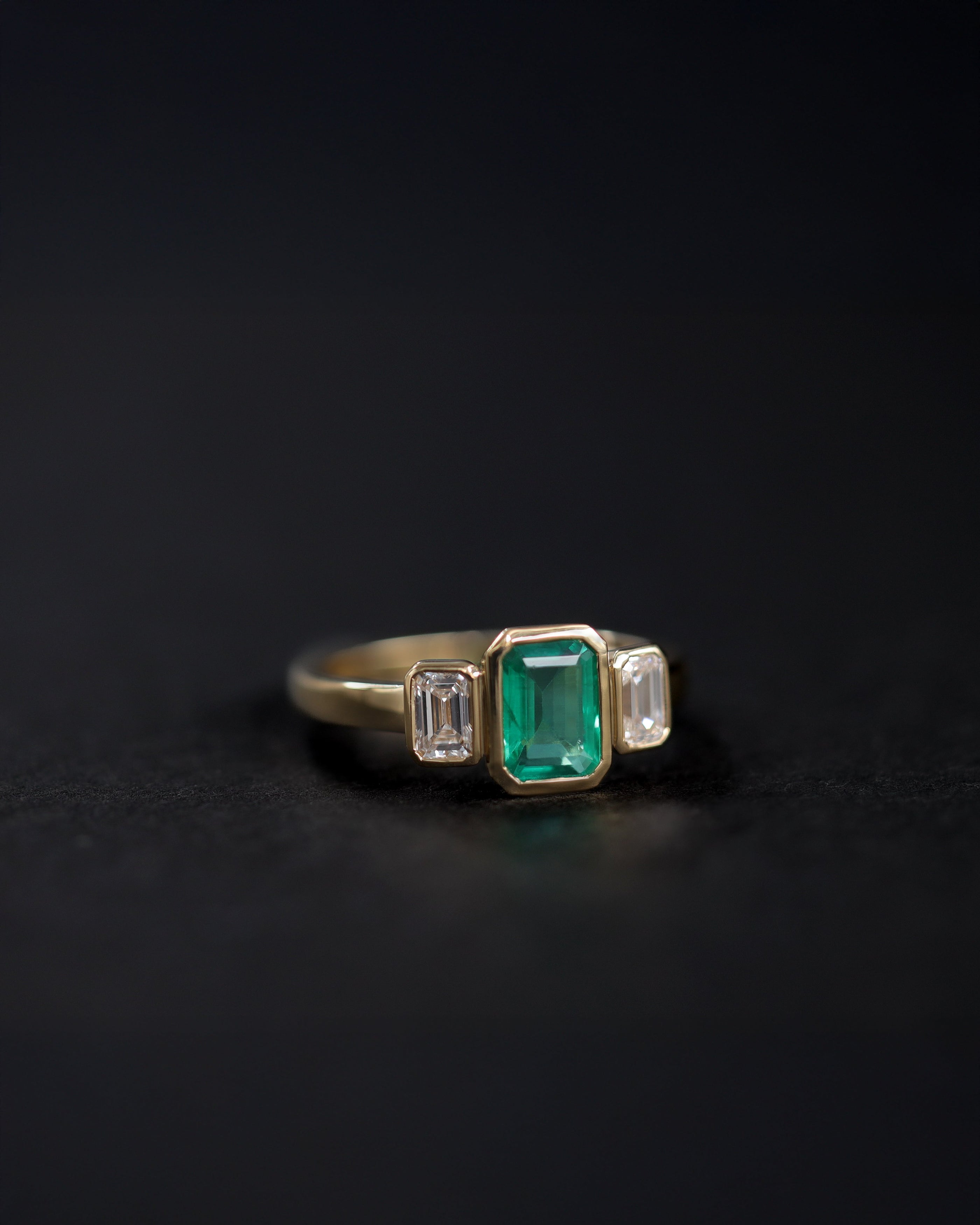 Bezel set 3 stone engagement ring with a vibrant emerald center stone, flanked by two emerald cut diamonds. Handmade in Auckland, New Zealand by award winning master jewellers 