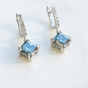 Pair of white gold drop earrings with square-cut blue diamonds and pavé diamond-set hooks displayed. Handmade by our master jewellers and diamonds set by our master diamond setters.