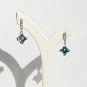 Pair of white gold drop earrings with square-cut blue diamonds and pavé diamond-set hooks displayed on a textured white stand.