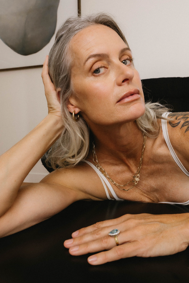 Woman with gray hair wearing a white tank top, gold necklace, and ring on a neutral background