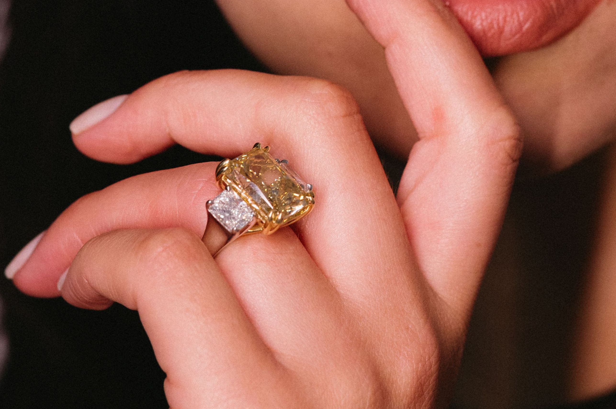 Close-up of a hand wearing a gold ring with a large yellow diamond and smaller diamonds on either side on a dark background